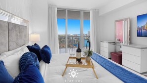Ett hotellrum med en säng, ett skrivbord, ett fönster med utsikt över staden och ett bricka med en flaska och glas.