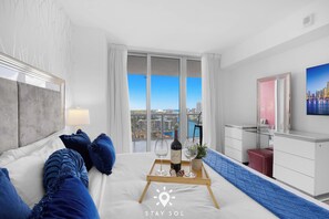 Ein Hotelzimmer mit Bett, Schreibtisch, Fenster mit Stadtblick und Tablett mit Flasche und Gläsern.