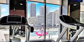 Un gimnasio con equipo de ejercicio y vistas a un paisaje urbano a través de amplias ventanas.