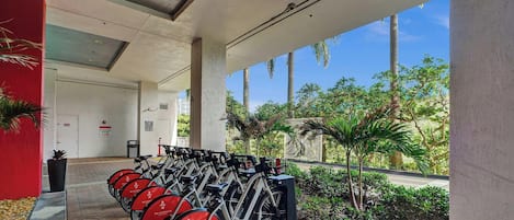 A covered outdoor area with a row of stationary bikes for rent, a pathway leading to a door marked 'PACKAGE ROOM,' and a view of palm trees and greenery.