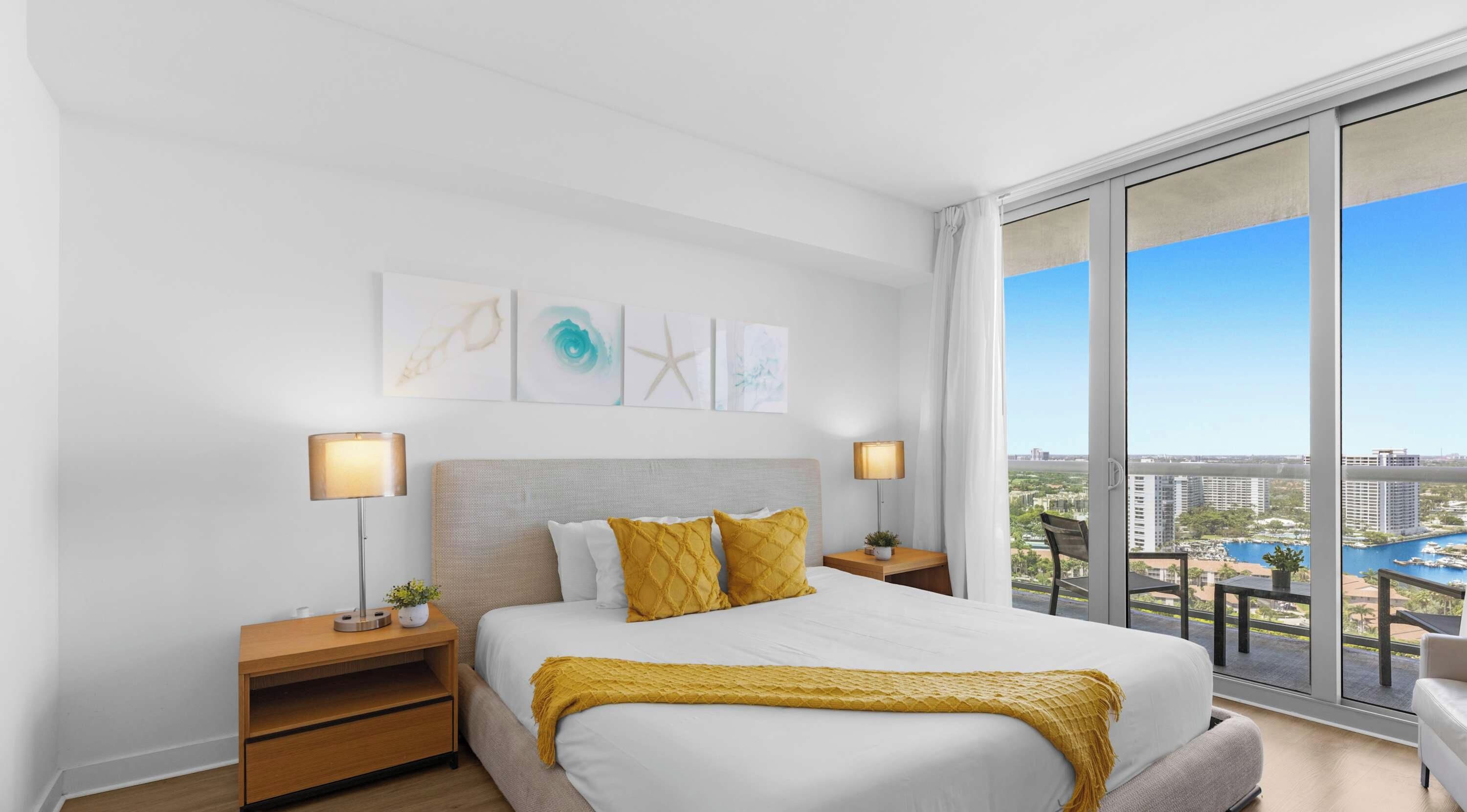 A hotel room with a large bed, two bedside tables, a view of the city, and artwork on the wall.
