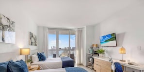 A modern hotel room with a bed, bedside table, lamp, and a view of the city through large windows. - High-Floor Studio | Pool & GYM (Hallandale Beach)