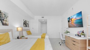 A modern hotel room with two beds, a desk, a chair, and a large window with a beach view. - Panoramic Marina View · 2BR · Beachwalk (Hallandale Beach)