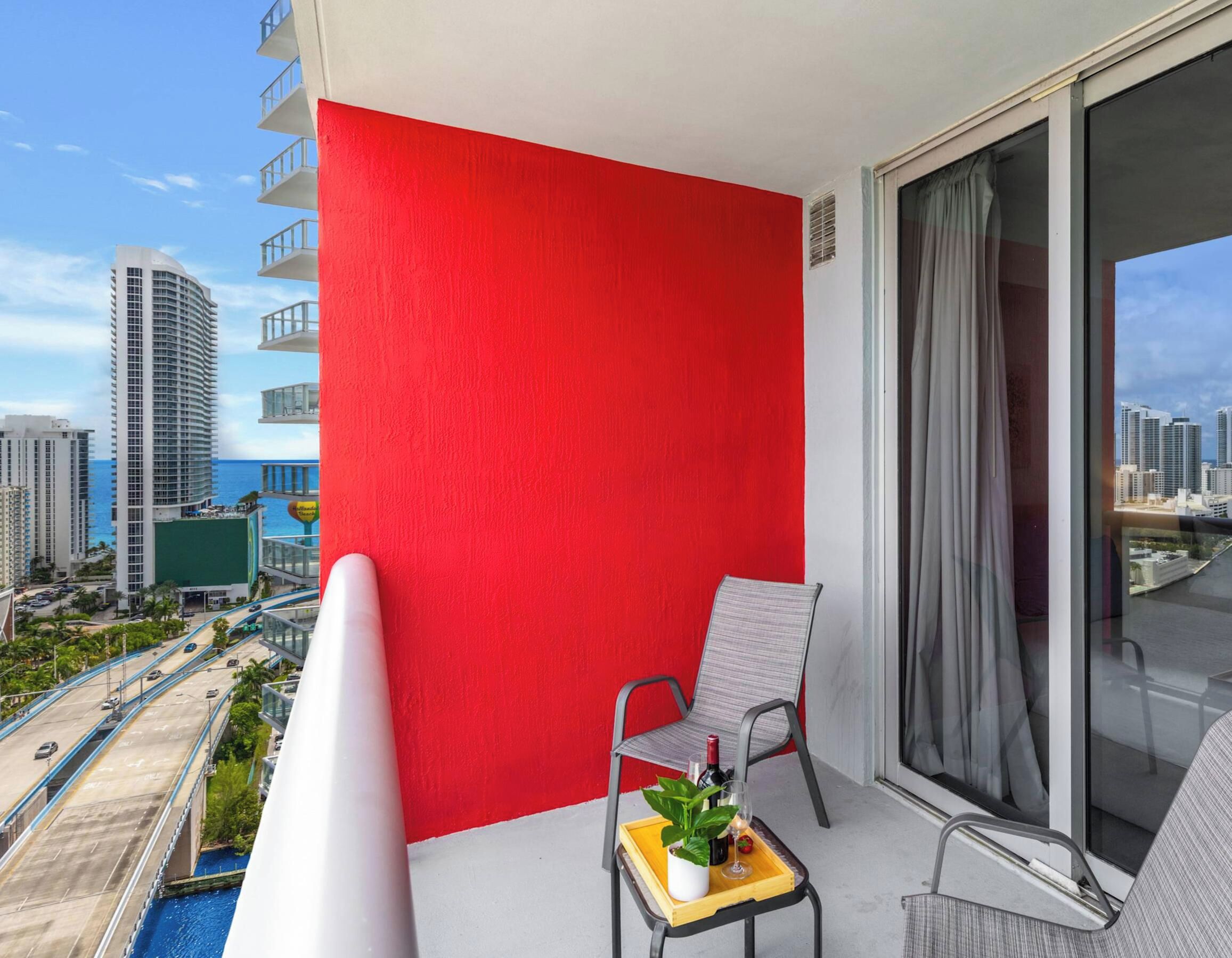 A balcony with a red wall, a chair, a small table with a plant and a bottle, and a view of a cityscape and water.