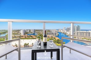Un balcon avec une table, deux chaises, une bouteille de vin et deux verres à vin. La vue comprend un paysage urbain et une étendue d’eau.