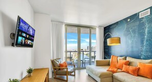 A modern hotel room with a large window, a flat-screen TV, a sofa, a chair, and a map wall. - Bay & City View 2BR · Beachwalk High Floor (Hallandale Beach)