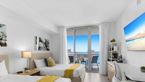 A hotel room with two beds, a desk, a chair, a balcony with a city view, and artwork on the walls. - Elevated Sunset View · 2BR Beachwalk Resort (Hallandale Beach)