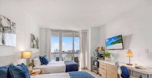 Ein modernes Hotelzimmer mit Bett, Nachttisch, Lampe und Blick auf die Stadt durch große Fenster.
