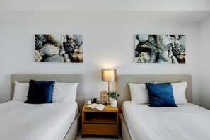 A hotel room with two beds, each with a white duvet and a blue pillow. There are two bedside tables with lamps, a small plant, and a book. The walls are adorned with abstract art. - Upper Studio | Bay & Sunny Isles Skyline (Hallandale Beach)