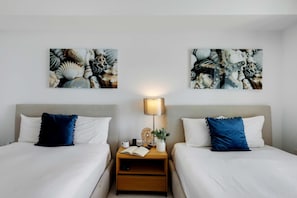 Ein Hotelzimmer mit zwei Betten, jeweils mit weißer Daunendecke und blauem Kopfkissen. Zwei Nachttische mit Lampen, eine kleine Pflanze und ein Buch sind ebenfalls vorhanden. Die Wände sind mit abstrakter Kunst geschmückt.