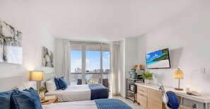 A modern hotel room with a bed, bedside table, lamp, and a view of the city through large windows. - Upper Studio | Intracoastal & Miami Skyline (Hallandale Beach)