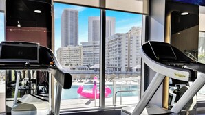 Ett gym med träningsutrustning och utsikt över en stadssiluett genom stora fönster.