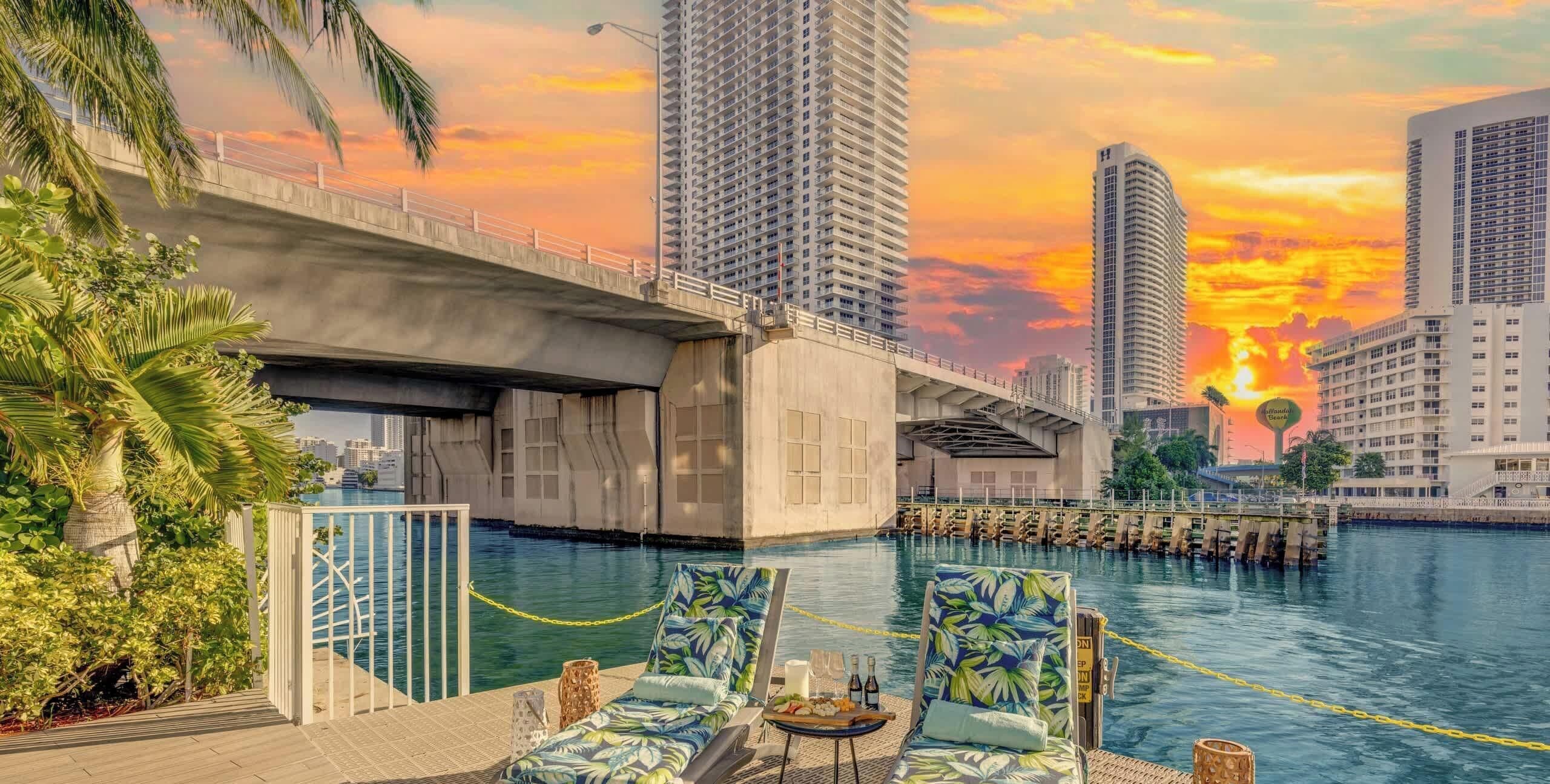 A waterfront pool area with lounge chairs, a dining table with drinks, and a view of high-rise buildings under a sunset sky.