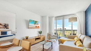 Un salon moderne avec une table basse en verre, un canapé, un coin repas comprenant une table et des chaises, et une grande fenêtre donnant sur la ville.
