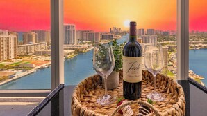 Una mesa con una botella de vino y dos copas, ubicada en un balcón con vistas a un paisaje urbano y un cuerpo de agua durante el atardecer.