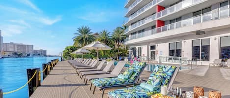 Zona de piscina con tumbonas, una mesa de comedor con aperitivos y bebidas, y vistas al agua y al horizonte de la ciudad.
