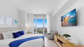 A hotel room with a large bed, a desk with a lamp, a chair, and a painting of a sunset. - New! 1BR Intracoastal & Marina View, Pool & Beach (Hallandale Beach)