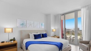 Habitación de hotel con una cama grande, dos mesitas de noche, una silla y un balcón con vista a la ciudad.