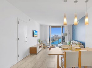 A modern living room with a dining area, large windows with a city view, and a minimalist decor. - Sunset View Apt | Full Kitchen & Beach Shuttle (Hallandale Beach)