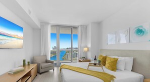 A modern hotel room with a large bed, a chair, a desk, and a view of a cityscape and water. - Sunset View Apt | Full Kitchen & Beach Shuttle (Hallandale Beach)