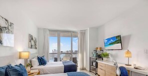 A modern hotel room with a bed, bedside table, lamp, and a view of the city through large windows. - New! High-Floor Studio Bay & Private Balcony (Hallandale Beach)