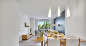 Ein moderner Essbereich mit einem Holztisch, gedeckt für eine Mahlzeit, einem an der Wand befestigten Fernseher und Blick ins Freie durch große Fenster.
