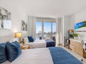Ein modernes Hotelzimmer mit Bett, Nachttisch, Lampe und Blick auf die Stadt durch große Fenster.