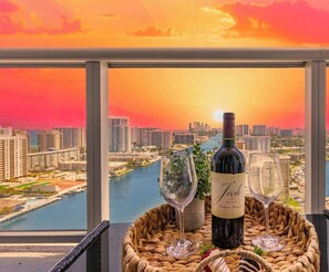A bottle of wine, two wine glasses, and a woven basket on a balcony with a cityscape and river view. - Sunset Panorama 2BR · Beachwalk High Floor (Hallandale Beach)