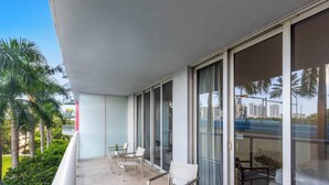 Un balcón con zona de estar, ventanas amplias y vistas a palmeras y edificios.