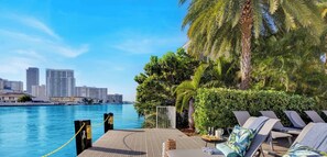 A waterfront balcony with lounge chairs, a pool, and a city skyline in the background. - Stylish 1BR Near the Beach  Resort Life (Hallandale Beach)