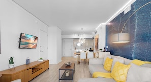 A modern living room with a sofa, a TV, and a dining area with a kitchen in the background. - Modern 1BR- APT - Resort‑Style Experience (Hallandale Beach)