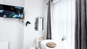 Ett hotellrum med en tv som är monterad på väggen, ett vitt bord med en vävd bordsduk, en vit stol, ett element och ett fönster med gardiner.