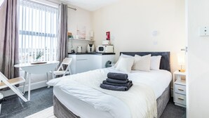 Ett hotellrum med en säng, ett skrivbord, en stol, ett litet pentry och ett fönster med gardiner.