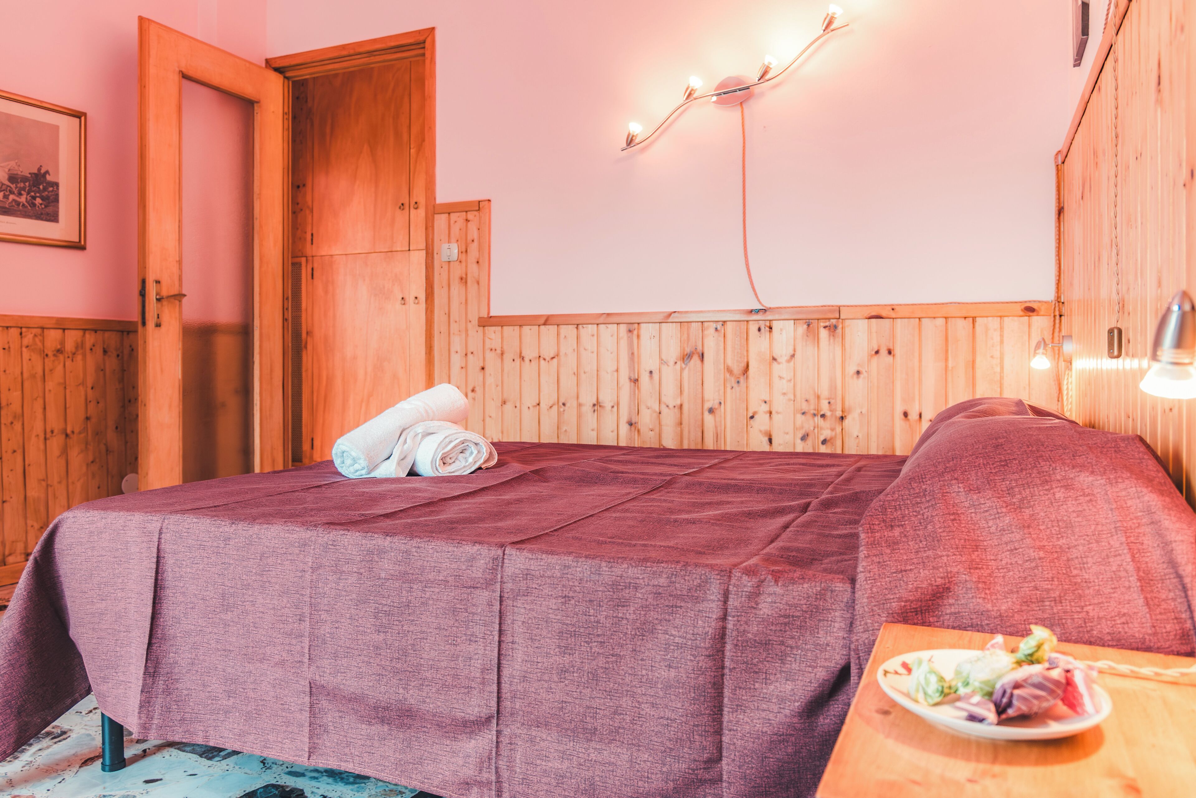 Una habitación con una cama, una puerta de madera, una lámpara de pared y una pequeña mesa con un plato de comida.