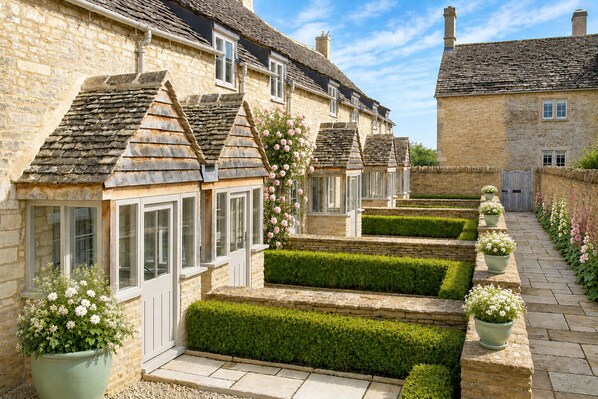 A stone building with multiple windows, a garden with neatly trimmed hedges, and a pathway lined with flower pots.