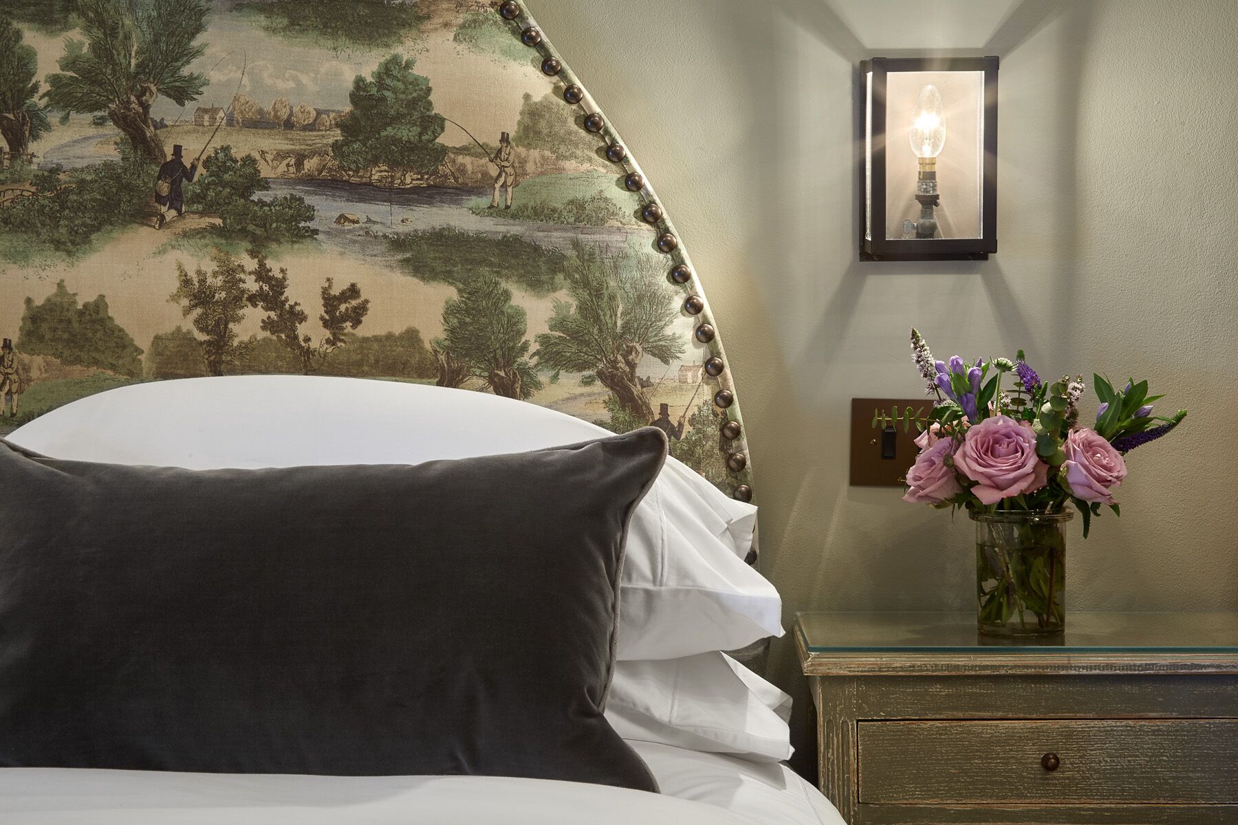 A bed with white and dark pillows, a nightstand with a vase of flowers, and a wall with a framed picture and a wall sconce.
