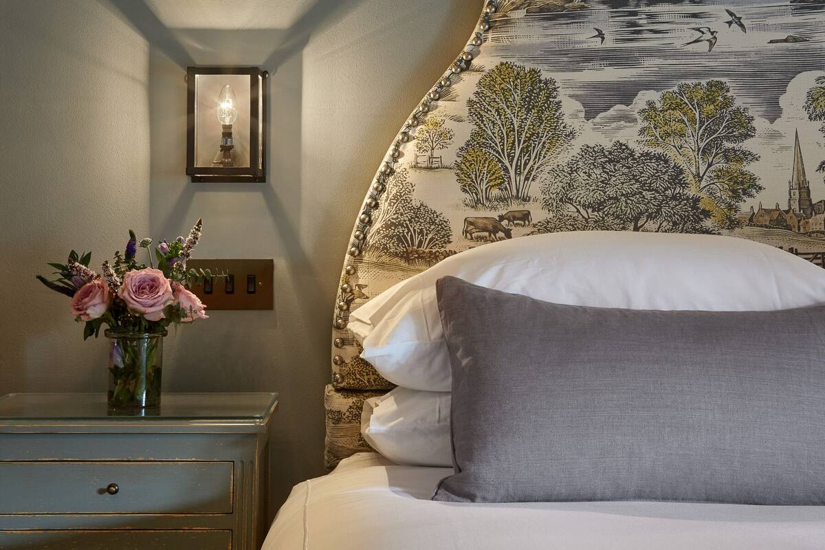 A bedroom with a bed, a nightstand with a vase of flowers, a wall-mounted light, and a patterned wallpaper.