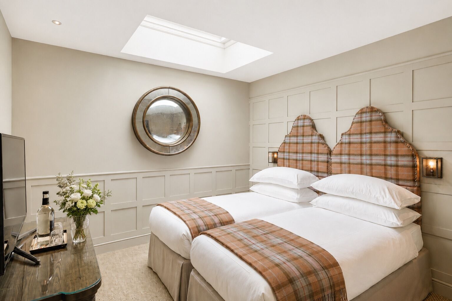 Une chambre d’hôtel avec un lit, un miroir rond, une table de chevet ornée d’un vase de fleurs et un téléviseur.