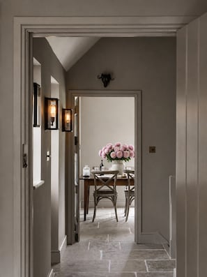 Un couloir mène à une salle à manger où une table est dressée pour deux personnes, agrémentée d’un vase de fleurs et éclairée par des appliques murales.