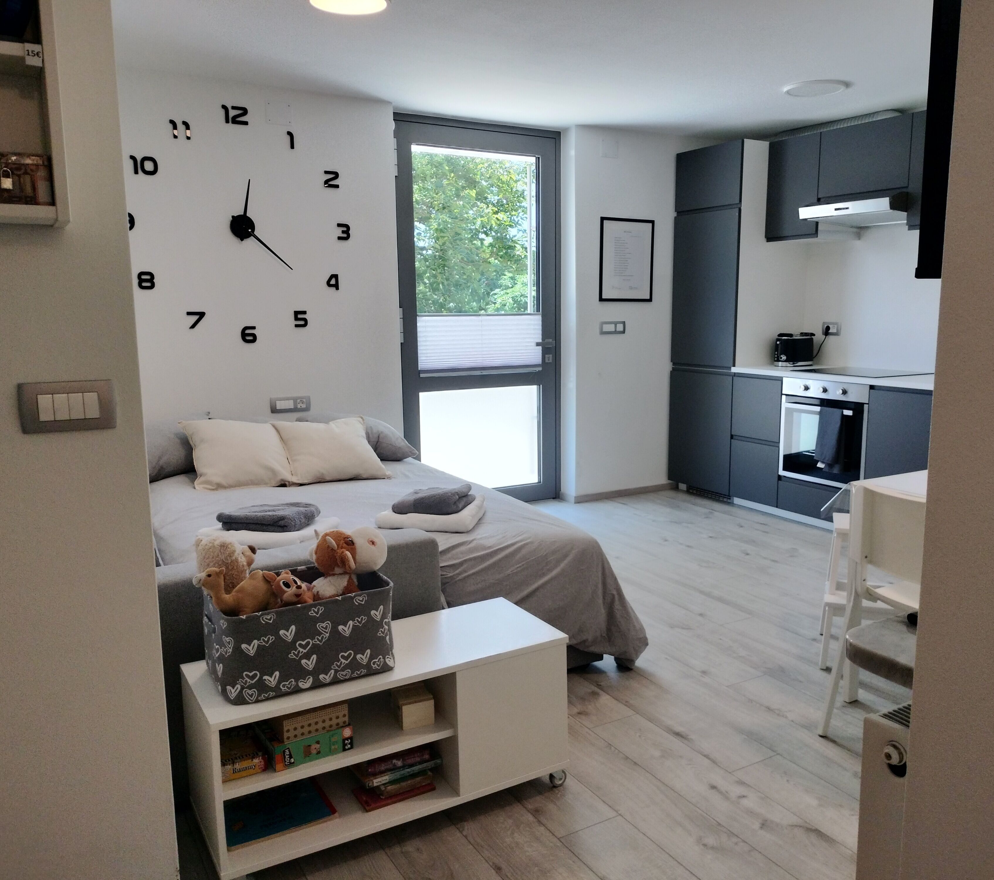 Ein modernes Schlafzimmer mit einer großen Wanduhr, einem Bett mit Kissen, einem Nachttisch mit Büchern, einem Fenster und einer Küchenzeile.