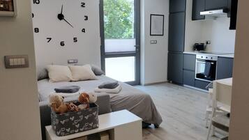 Ein modernes Schlafzimmer mit einer großen Wanduhr, einem Bett mit Kissen, einem Nachttisch mit Büchern, einem Fenster und einer Küchenzeile.