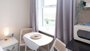 A small, bright room with a window, a white table, two wicker chairs, a bedside table with a lamp, and a microwave. - Bayview Aparthotel by Guestz (Morecambe)