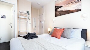 A hotel room with a bed, a nightstand, a wall-mounted shelf with plants, a door, and a painting. - Bayview Aparthotel by Guestz (Morecambe)