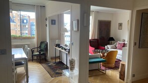Een moderne woonkamer met een eethoek, een rode fauteuil, een gele fauteuil, een witte bank en uitzicht op gebouwen door grote ramen.