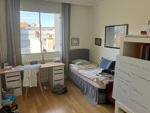 Une chambre à coucher comprenant un lit, un bureau, un radiateur, une fenêtre avec des rideaux et un tableau blanc sur lequel sont écrites des équations chimiques.