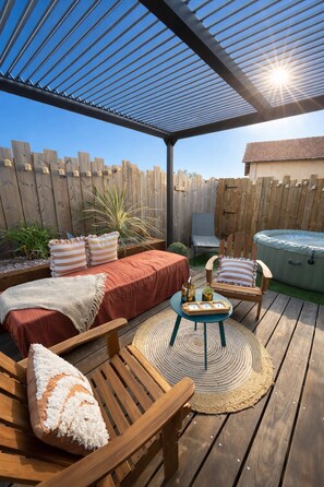 Eine überdachte Terrasse mit Holzdeck, einer gepolsterten Sitzgruppe, einem kleinen Tisch und einem Whirlpool.