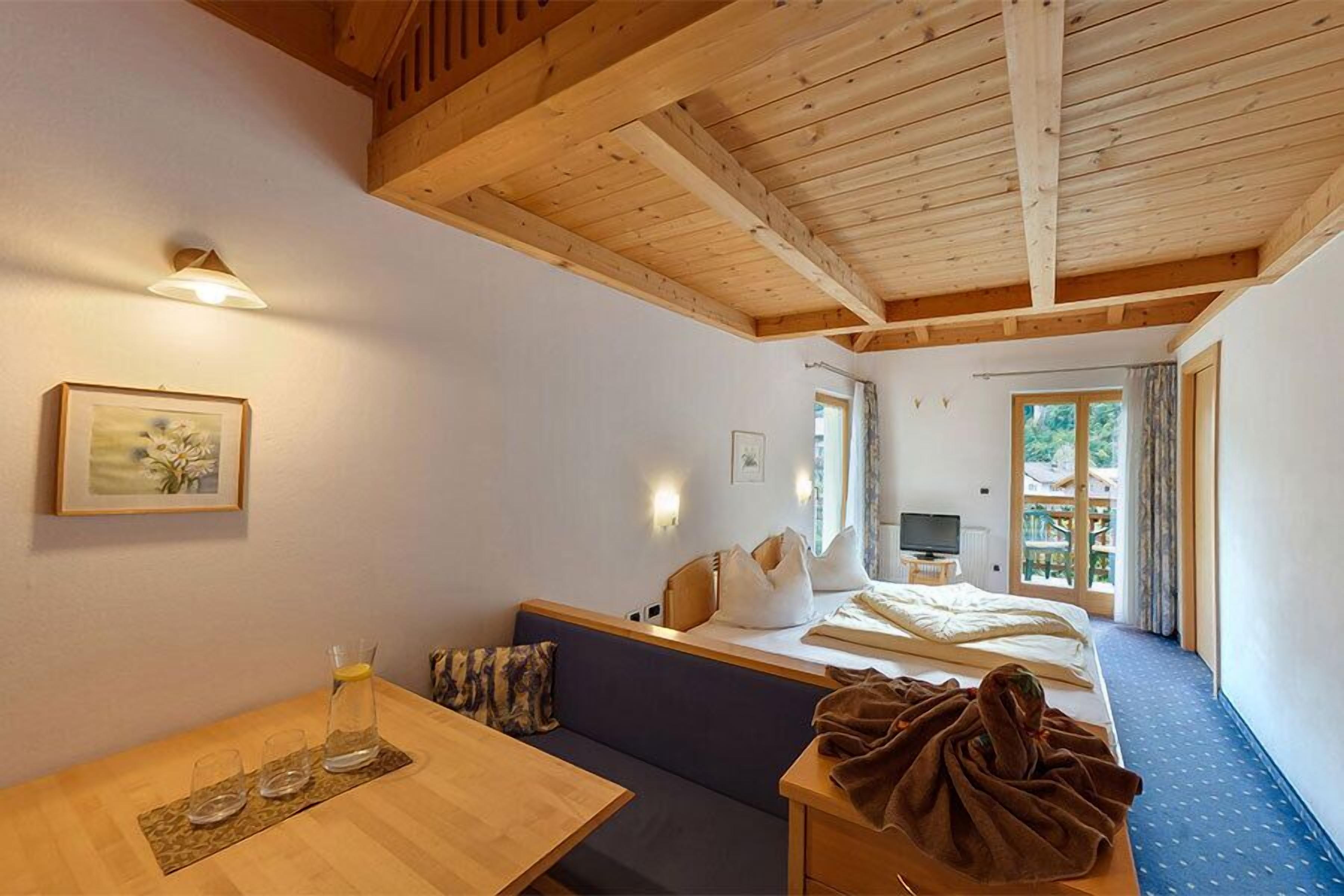 A room with a wooden ceiling, a bed with pillows, a table with glasses and a bottle, and a television on a stand.