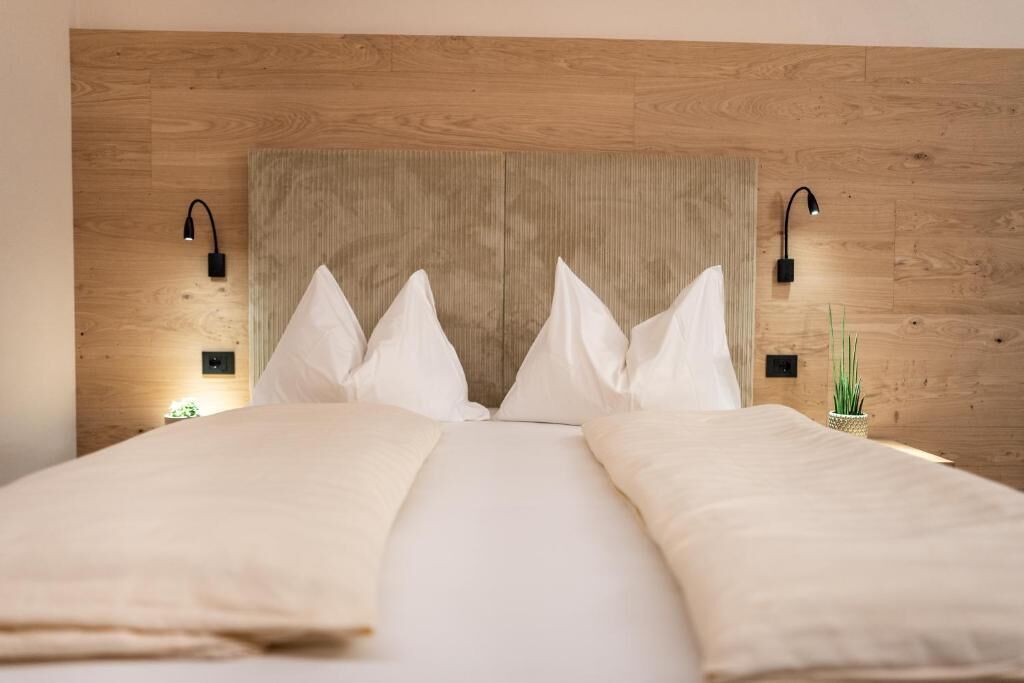 Una cama con ropa blanca, cabecera de madera, dos lámparas de noche y una pequeña planta en maceta.