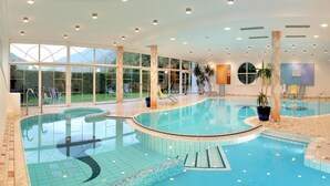 An indoor swimming pool with clear glass walls, lounge chairs, and potted plants.