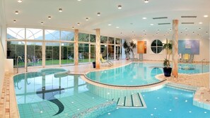 An indoor swimming pool with clear glass walls, lounge chairs, and potted plants.